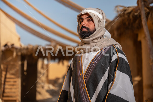 قرية تاريخية تراثية، يوم بدينا22 فبراير, ذكرى تأسيس الدولة السعودية الأولى1727م, ارتداء الزي الشعبي في مناسبة وطنية, صورة مقربة لرجل عربي خليجي سعودي يرتدي البشت والغترة يقف أمام بيت طيني قديم وينظر للأعلى بايماءات الفخر، خلفية سقف المنزل المغطى بسعف النخيل