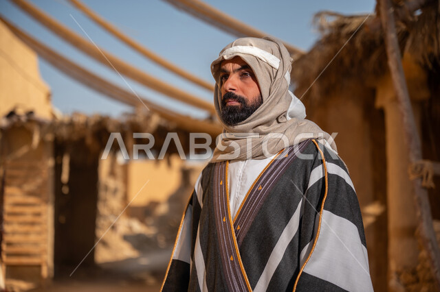 بلدة تاريخية تراثية، يوم بدينا22 فبراير, ذكرى تأسيس الدولة السعودية الأولى1727م, ارتداء الزي الشعبي في مناسبة وطنية, صورة مقربة لرجل عربي خليجي سعودي يرتدي البشت والغترة يقف أمام بيت طيني قديم وينظر للأعلى بايماءات الفخر، خلفية سقف المنزل المغطى بسعف النخيل