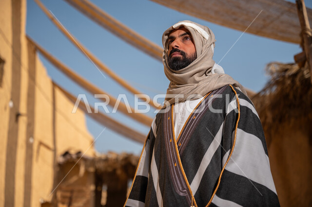 يوم بدينا22 فبراير, ذكرى تأسيس الدولة السعودية الأولى1727م, ارتداء الزي الشعبي في مناسبة وطنية, قرية تاريخية تراثية، صورة مقربة لرجل عربي خليجي سعودي يرتدي البشت والغترة يقف أمام بيت طيني قديم وينظر  للأعلى بايماءات الفخر، مظلات قماشية ممتدة بين البيوت