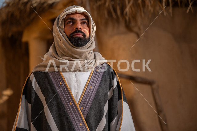بلدة تراثية تاريخية، ذكرى تأسيس الدولة السعودية الأولى1727م,  ارتداء الزي الشعبي لإحياء عيد ومناسبة وطنية, يوم بدينا22 فبراير, رجل عربي خليجي سعودي يرتدي البشت والغترة يقف أمام بيت طيني قديم وينظر للأعلى بايماءات التأمل والفخر، خلفية السقف المغطى بسعف النخل