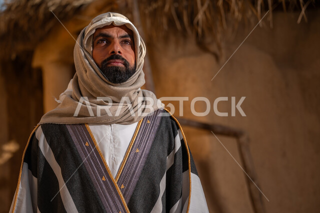 بلدة تراثية تاريخية، ذكرى تأسيس الدولة السعودية الأولى1727م,  ارتداء الزي الشعبي لإحياء عيد ومناسبة وطنية, يوم بدينا22 فبراير, رجل عربي خليجي سعودي يرتدي البشت والغترة يقف أمام بيت طيني قديم وينظر للأعلى بايماءات التأمل والفخر، خلفية السقف المغطى بسعف النخل