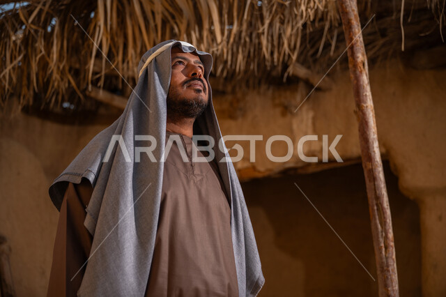 يوم بدينا22 فبراير, ذكرى تأسيس الدولة السعودية الأولى1727م, ارتداء الزي الشعبي في مناسبة وطنية, قرية تاريخية تراثية، رجل عربي خليجي سعودي يرتدي الثوب التقليدي والغترة يقف أمام بيت طيني قديم وينظر إلى الأعلى بايماءات الفخر، خلفية سقف المنزل المغطى بسعف النخل