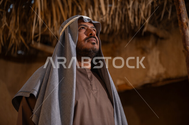 ذكرى تأسيس الدولة السعودية الأولى1727م, ارتداء الزي الشعبي في مناسبة وطنية, قرية تاريخية تراثية، يوم بدينا22 فبراير, رجل عربي خليجي سعودي يرتدي الثوب التقليدي والغترة يقف أمام بيت طيني قديم وينظر إلى الأعلى بايماءات الفخر، خلفية سقف المنزل المغطى بسعف النخل