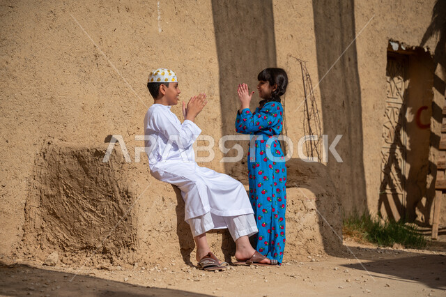 ألعاب متوارثة من الأجداد عبر الأجيال، قضاء وقت ممتع مع الأخ، بنت عربية خيليجية سعودية ترتدي فستان أزرق تقوم باللعب والمرح برفقة أخيها بإيماءات السعادة والسرور، إحياء ذكرى تأسيس الدولة السعودية الأولى 1727 م, يوم بدينا 22 فبراير, منزل طيني قديم في قرية تاريخية