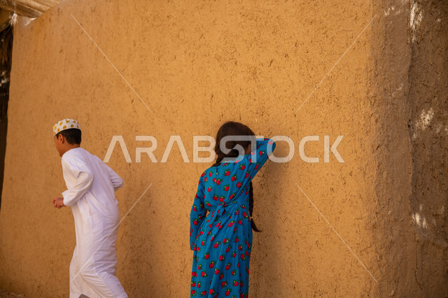 Games passed down through generations, commemorating the founding of the first Saudi state in 1727 AD, the day we started on February 22, an old clay house in a historical archaeological village, a boy and his sister, two Saudi Gulf Arabs, wearing traditional folk costumes and playing a game of seek and hide, having fun and enjoying the time