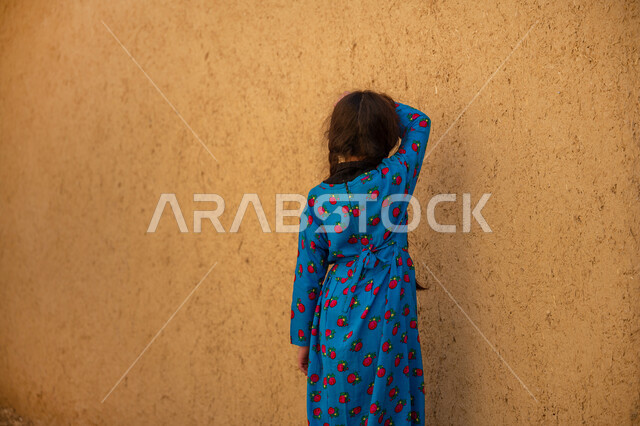 Games passed down through generations, commemorating the founding of the first Saudi state in 1727 AD, the day we started on February 22, an old clay house in a historical archaeological village, a Saudi Arabian Gulf girl wearing a traditional blue folk dress closes her eyes and plays hide and seek