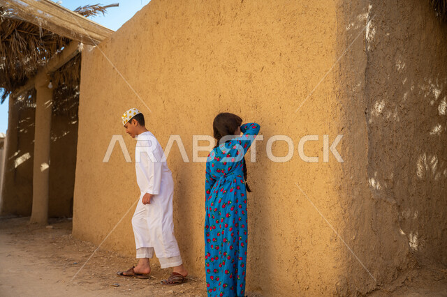 اللهو والمرح والاستمتاع بالوقت، ألعاب متوارثة عبر الأجيال، إحياء ذكرى تأسيس الدولة السعودية الأولى 1727 م, يوم بدينا 22 فبراير, منزل طيني قديم في قرية تاريخية أثرية، ولد وأخته عربيان خليجيان سعوديان يرتديان الزي الشعبي التقليدي ويلعبان لعبة البحث والاختباء
