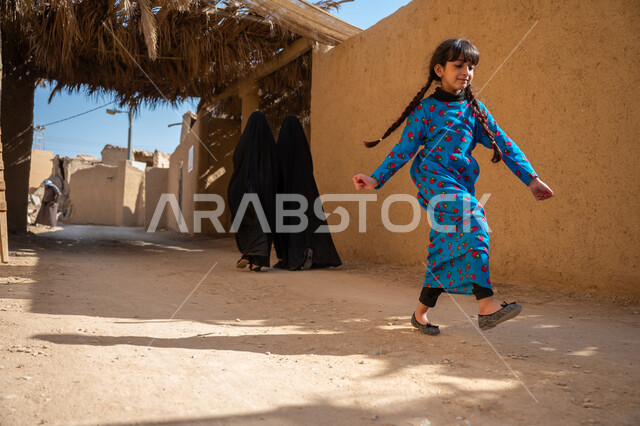 يوم بدينا 22 فبراير, منزل طيني قديم في قرية تاريخية أثرية، إحياء ذكرى تأسيس الدولة السعودية الأولى 1727 م, فتاة عربية خليجية سعودية ترتدي فستان أزرق شعبي تقليدي تقوم بالقفز واللعب في الحي بإيماءات السعادة والسرور