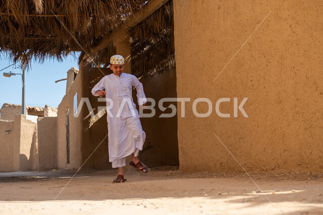منزل طيني قديم في قرية تاريخية أثرية، إحياء ذكرى تأسيس الدولة السعودية الأولى 1727 م, يوم بدينا 22 فبراير, صبي عربي خليجي سعودي يرتدي الثوب التقليدي والقبعة يقوم بالقفز واللعب بإيماءات السعادة والسرور