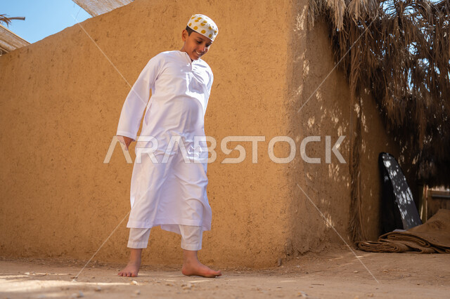 اللعب والاستمتاع في قرية تاريخية قديمة، إحياء ذكرى تأسيس الدولة السعودية الأولى1727م, يوم بدينا 22 فبراير, صبي عربي خليجي سعودي يرتدي الثوب التقليدي والقبعة حافي القدمين يقوم باللهو والمرح أمام منزل طيني قديم