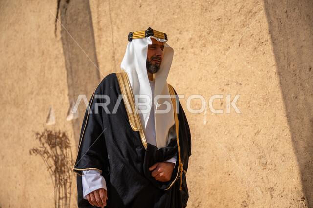 إحياء ذكرى تأسيس الدولة السعودية الأولى 1727 م, يوم بدينا 22 فبراير, رجل عربي خليجي سعودي يرتدي البشت والغترة والعقال الذهبي العريض يقف أمام منزل طيني قديم وينظر إلى مكان ما بإيماءات التأمل، قرية تاريخية قديمة، ارتداء الزي الشعبي  التقليدي في عيد ومناسبة وطنية