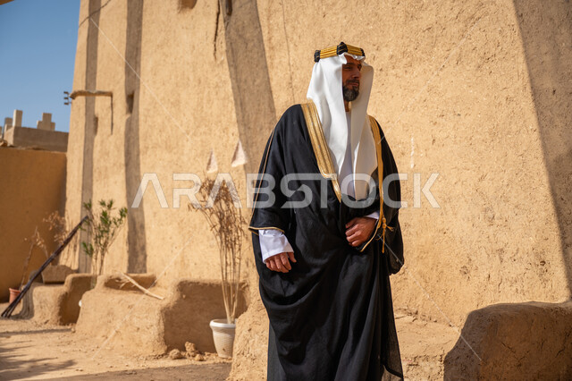 ارتداء الزي الشعبي  التقليدي في عيد ومناسبة وطنية، إحياء ذكرى تأسيس الدولة السعودية الأولى 1727 م, يوم بدينا 22 فبراير, رجل عربي خليجي سعودي يرتدي البشت والغترة والعقال الذهبي العريض يقف أمام منزل طيني قديم وينظر إلى مكان ما بإيماءات التأمل، قرية تاريخية قديمة