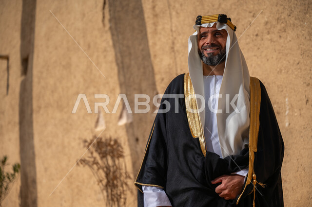 إحياء ذكرى تأسيس الدولة السعودية الأولى 1727 م, يوم بدينا 22 فبراير, رجل عربي خليجي سعودي يرتدي البشت والغترة والعقال الذهبي العريض يقف أمام منزل طيني قديم وينظر إلى مكان ما بإيماءات التأمل، قرية تاريخية قديمة، ارتداء الزي الشعبي  التقليدي في عيد ومناسبة وطنية