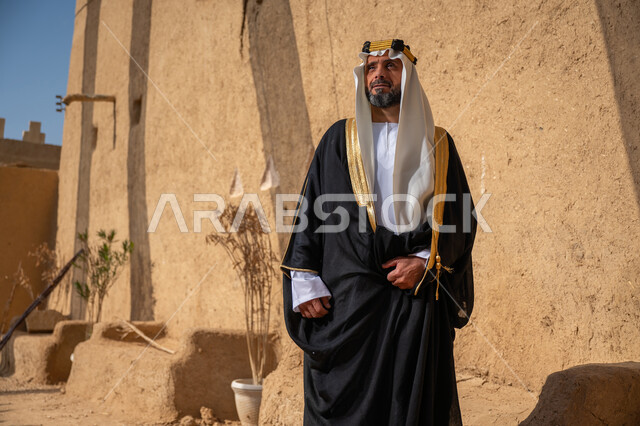 الشموخ والاعتزاز والفخر، رجل عربي خليجي سعودي يرتدي الغترة والعقال الذهبي والبشت يقف أمام جدار طيني قديم, يوم بدينا 22 فبراير، ذكرى تأسيس الدولة السعودية الأولى في 1727م، ارتداء الزي الشعبي في المناسبة الوطنية، إظهار حب الوطن والانتماء إليه