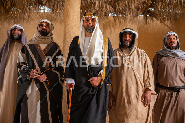 يوم بدينا 22 فبراير، إيماءات الثقة والفخر بإنجازات المملكة، ذكرى تأسيس الدولة السعودية الأولى 1727م، إظهار الولاء والحب للوطن, مجموعة رجال عرب خليجين سعوديين يرتدون الزي الشعبي التقليدي في المناسبة الوطنية يقفون في بيت من الطين ينظرون إلى الكاميرا بتعابير الشموخ