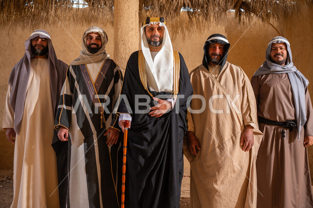 يوم بدينا 22 فبراير، إيماءات الثقة والفخر بإنجازات المملكة، ذكرى تأسيس الدولة السعودية الأولى 1727م، إظهار الولاء والحب للوطن, مجموعة رجال عرب خليجين سعوديين يرتدون الزي الشعبي التقليدي في المناسبة الوطنية يقفون في بيت من الطين ينظرون إلى الكاميرا بتعابير الشموخ