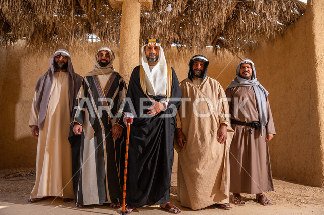 إيماءات الثقة والفخر بإنجازات المملكة، ذكرى تأسيس الدولة السعودية الأولى 1727م، إظهار الولاء والحب للوطن, مجموعة رجال عرب خليجين سعوديين يرتدون الزي الشعبي التقليدي في المناسبة الوطنية يقفون في بيت من الطين ينظرون إلى الكاميرا بتعابير الشموخ، يوم بدينا 22 فبراير