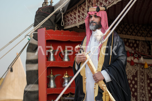 Passion and love for owning traditional swords, standing in front of a popular tent with gestures of pride and honor, a Saudi Arabian Gulf man wearing the traditional bisht and shemagh holding a golden sword in his hand, showing off the traditional Saudi weapon, the day we started, February 22, the anniversary of the founding of the first Saudi state 1727 AD
