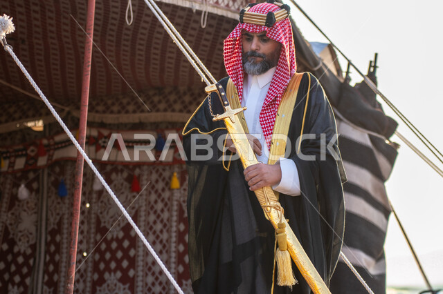 حب وشغف امتلاك السيوف التراثية, الوقوف أمام خيمة شعبية بإيماءات الاعتزاز والفخر, رجل عربي خليجي سعودي يرتدي البشت و الشماغ التقليدي يحمل بيده السيف الذهبي, التباهي بالسلاح التقليدي السعودي, يوم بدينا 22 فبراير ، ذكرى تاسيس الدوله السعوديه الاولى 1727م