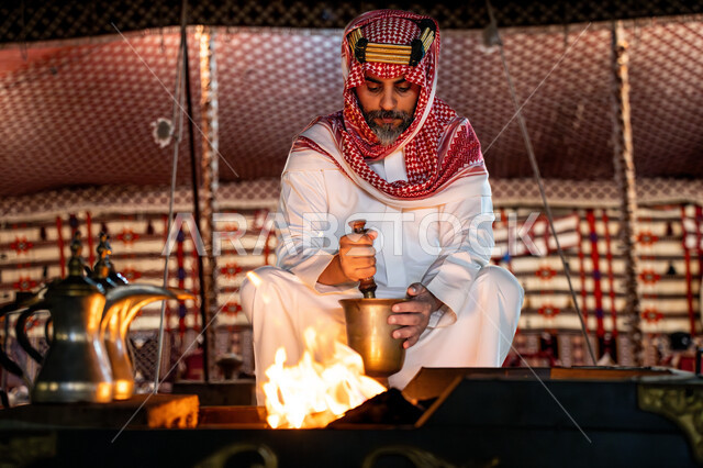 أسلوب الحياة القديمه في الباديه، طحن حب البن في النجر النحاسي, يوم بدينا 22 فبراير، ذكرى تاسيس الدوله السعوديه الاولى 1727م, رجل عربي خليجي سعودي يرتدي الثوب التقليدي و الشماغ يجلس أمام شعلة النار، اجواء شتوية ليلية