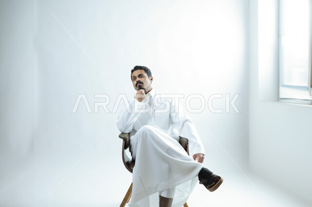 مفهوم التفكير والتخطيط واتخاذ القرار, بورتريه لرجل عربي خليجي يرتدي الثوب الابيض يجلس على كرسي خشبي في استوديو أبيض يضع يده على ذقنه بإيماءات التأمل في المستقبل, حالة التفكير العميق, خلفية بيضاء