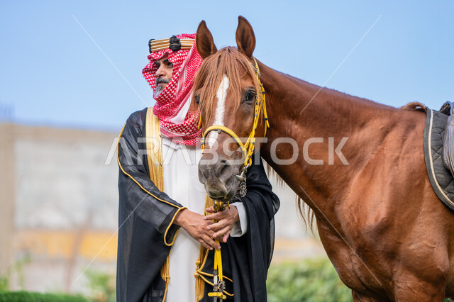 ذكرى تأسيس الدولة السعودية الأولى في 1727م, النظر الى شيء ما بتعابير الفخر, توراث حب وتربية الجياد من الأجداد, الزي الشعبي في المناسبة الوطنية، يوم بدينا 22 فبراير, رعاية الخيل والاهتمام به، رجل عربي خليجي سعودي يرتدي البشت و الشماغ يقف بجانب فرسه البني الأصيل