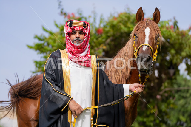 النظر الى الكاميرا بتعابير الفخر, توراث حب وتربية الجياد من الأجداد, الزي الشعبي في المناسبة الوطنية، يوم بدينا 22 فبراير, ذكرى تأسيس الدولة السعودية الأولى في 1727م, رعاية الخيل والاهتمام به، رجل عربي خليجي سعودي يرتدي البشت و الشماغ يقف بجانب فرسه البني الأصيل