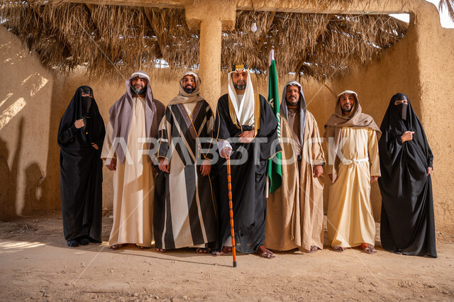ذكرى تأسيس الدولة الأولى 1727م، يوم بدينا 22 فبراير, علم المملكة العربية السعودية، إظهار الولاء والحب للوطن, مجموعة رجال ونساء عرب خليجين سعوديين يرتدون الزي الشعبي التقليدي في المناسبة الوطنية يقفون أمام بيت طيني قديم ينظرون إلى الكاميرا بإيماءات الشموخ والاعتزاز