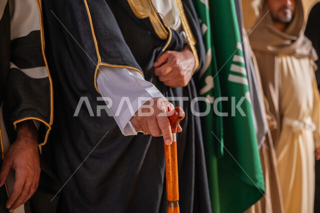 حب الوطن والانتماء إليه, صورة مقربة لرجل عربي خليجي سعودي يرتدي البشت يمسك بيده العكاز يقف بجانب علم المملكة العربية السعودية، ارتداء الزي الشعبي في المناسبة الوطنية، ذكرى تأسيس الدولة السعودية الأولى 1727م، يوم بدينا 22 فبراير، الفخر والاعتزاز بإنجازات البلاد