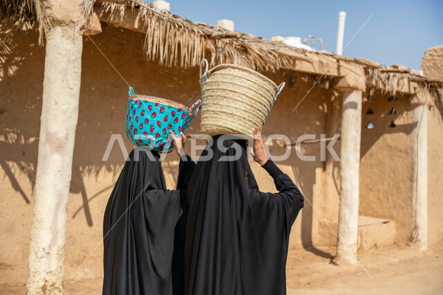 المشي في الاحياء والمنازل الطينية، صورة من الخلف لسيدتين عربيتين خليجيتين سعوديتين ترتديان العباءة والحجاب تحملان سلة من القش على رأسهما, منتج محلي وطني، مهنة تقليدية قديمة, يوم بدينا  22 فبراير، ذكرى تأسيس الدولة السعودية الأولى في 1727م