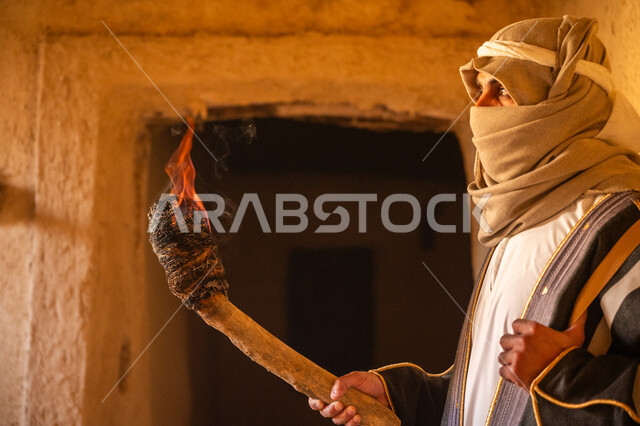 الحفاظ على تاريخ وتراث المملكة، القتال والحرب بالبنادق للحماية وتأسيس الدولة السعودية الأولى 1727م، صورة مقربة لرجل عربي خليجي سعودي يرتدي الزي الشعبي يحمل البندقية وشعلة النار يقف في بيت طيني، يوم بدينا 22 فبراير، الحراسة والمراقبة للدفاع عن الوطن