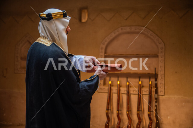 مجموعة من البنادق المعلقة على جدار طيني، صورة من الجانب لرجل عربي خليجي سعودي يرتدي الغترة والعقال الذهبي والبشت يقوم بفتح مخطوطة جلد تاريخية، ارتداء الزي الشعبي في المناسبة الوطنية، يوم بدينا 22 فبراير, الخطابات والمراسلات التراثية لتأسيس الدولة السعودية الأولى 1727م