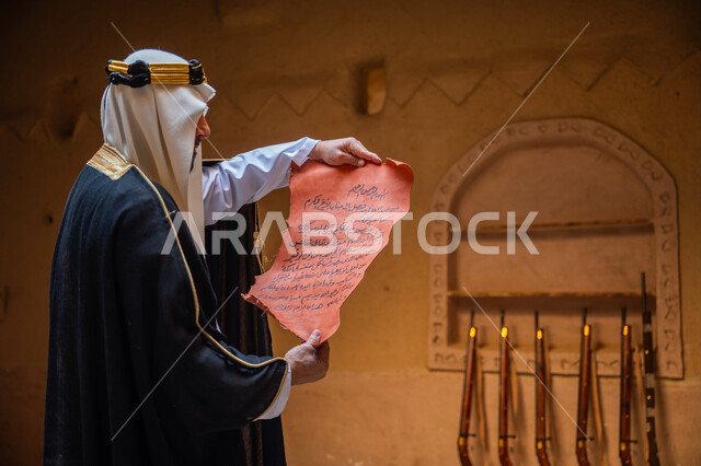 مجموعة من البنادق المعلقة على جدار طيني، صورة من الجانب لرجل عربي خليجي سعودي يرتدي الغترة والعقال الذهبي والبشت يقوم بقراءة مخطوطة جلد تاريخية، ارتداء الزي الشعبي في المناسبة الوطنية، يوم بدينا 22 فبراير, الخطابات والمراسلات التراثية لتأسيس الدولة السعودية الأولى 1727م