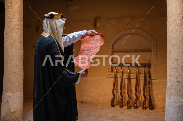 ارتداء الزي الشعبي في المناسبة الوطنية، مجموعة من البنادق المعلقة على جدار طيني، صورة من الجانب لرجل عربي خليجي سعودي يرتدي الغترة والعقال الذهبي والبشت يقوم بقراءة مخطوطة جلد تاريخية، يوم بدينا 22 فبراير, الخطابات والمراسلات التراثية لتأسيس الدولة السعودية الأولى 1727م