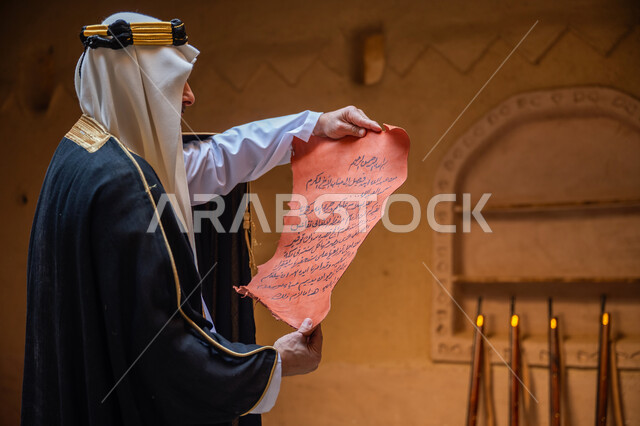 مجموعة من البنادق المعلقة على جدار طيني، صورة من الجانب لرجل عربي خليجي سعودي يرتدي الغترة والعقال الذهبي والبشت يقوم بقراءة مخطوطة جلد تاريخية، ارتداء الزي الشعبي في المناسبة الوطنية، يوم بدينا 22 فبراير, الخطابات والمراسلات التراثية لتأسيس الدولة السعودية الأولى 1727م