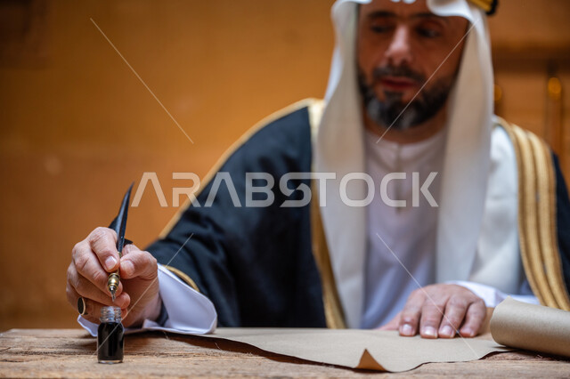 الخطابات والمراسلات التراثية لتأسيس الدولة السعودية الأولى 1727م، الفخر بأصالة المملكة، ارتداء الزي الشعبي في المناسبة الوطنية، صورة مقربة لرجل عربي خليجي سعودي يرتدي الغترة والعقال الذهبي والبشت يمسك بيده الريشة يقوم بالكتابة على مخطوطة جلد تاريخية، يوم بدينا 22 فبراير
