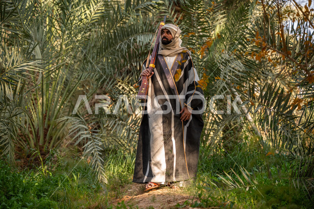 حرفة شعبية تراثية, اصطياد الحيوانات البرية في المزرعة, ذكرى يوم التأسيس السعودي 1727 م, يوم بدينا 22 فبراير, حمل السلاح للدفاع عن الوطن, رجل عربي خليجي سعودي يرتدي الزي التقليدي يحمل بيده بندقية ويسير بين أشجار النخيل, توارث مهنة الصيد