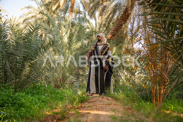 حمل السلاح للدفاع عن الوطن, رجل عربي خليجي سعودي يرتدي الزي التقليدي يحمل بيده بندقية ويسير بين أشجار النخيل, توارث مهنة الصيد, حرفة شعبية تراثية, اصطياد الحيوانات البرية في المزرعة, ذكرى يوم التأسيس السعودي 1727 م, يوم بدينا 22 فبراير
