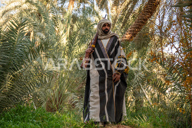 يوم بدينا 22 فبراير, حمل السلاح للدفاع عن الوطن, رجل عربي خليجي سعودي يرتدي الزي التقليدي يحمل بيده بندقية ويسير بين أشجار النخيل, توارث مهنة الصيد, حرفة شعبية تراثية, اصطياد الحيوانات البرية في المزرعة, ذكرى يوم التأسيس السعودي 1727 م