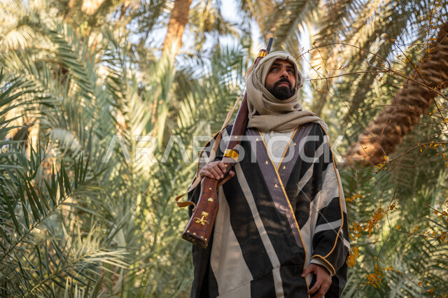 ذكرى يوم التأسيس السعودي 1727 م, يوم بدينا 22 فبراير, حمل السلاح للدفاع عن الوطن, رجل عربي خليجي سعودي يرتدي الزي التقليدي يحمل بيده بندقية ويسير بين أشجار النخيل, توارث مهنة الصيد, حرفة شعبية تراثية, اصطياد الحيوانات البرية في المزرعة