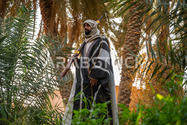 اصطياد الحيوانات البرية في المزرعة, ذكرى يوم التأسيس السعودي 1727 م, يوم بدينا 22 فبراير, حمل السلاح للدفاع عن الوطن, رجل عربي خليجي سعودي يرتدي الزي التقليدي يحمل بيده بندقية ويسير بين أشجار النخيل, توارث مهنة الصيد, حرفة شعبية تراثية