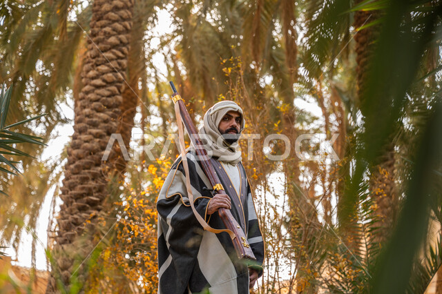 حرفة شعبية تراثية, اصطياد الحيوانات البرية في المزرعة, ذكرى يوم التأسيس السعودي 1727 م, يوم بدينا 22 فبراير, حمل السلاح للدفاع عن الوطن, رجل عربي خليجي سعودي يرتدي الزي التقليدي يحمل بيده بندقية ويسير بين أشجار النخيل, توارث مهنة الصيد