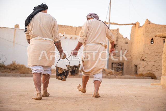 يوم بدينا 22 فبراير, الفخر والاعتزاز بالتراث السعودي العريق, تشييد وبناء البيوت الطينية التراثية التاريخية, إحياء ذكرى يوم التأسيس1727 م, بناء الدولة السعودية الأولى, صورة من الخلف لعاملان عربيان يقومان بنقل الطين ومواد البناء