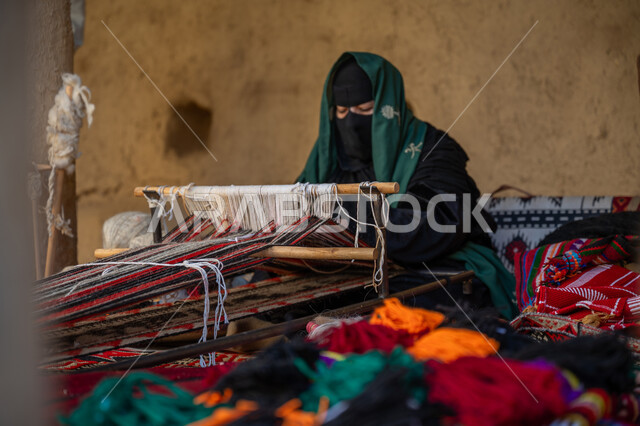 حرفة يدوية تاريخية, مهنة تقليدية قديمة, يوم بدينا 22 فبراير، ارتداء الزي الشعبي في المناسبة الوطنية، امرأة عربية خليجية سعودية ترتدي العباءة والبرقع تجلس في بيت من الطين وتقوم بحياكة وغزل الأنسجة والقماش, منتج محلي وطني، ذكرى تأسيس الدولة السعودية الأولى في 1727م