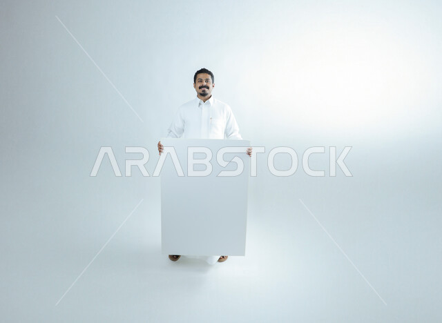 عرض موك أب مربع فارغ كبير، بورتريه لشاب عربي سعودي خليجي مبتسم يرتدي الثوب التقليدي يقف باستقامة ويحمل بيده لوحة بيضاء فارغة، استخدام لوح توضيحي في مجال التعليم والعروض الترويجية، الطول الكامل للجسم، خلفية بيضاء