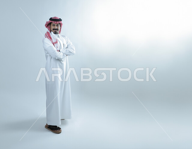 النظر إلى الكاميرا بايماءات الثقة بالنفس، تعابير البشاشة والسرور، مفهوم الاناقة والرجولة، الإهتمام بالمظهر الخارجي، بورتريه لرجل عربي خليجي سعودي يرتدي الثوب التقليدي والشماغ يقف بايدي متقاطعة، الطول الكامل للجسم، خلفية بيضاء