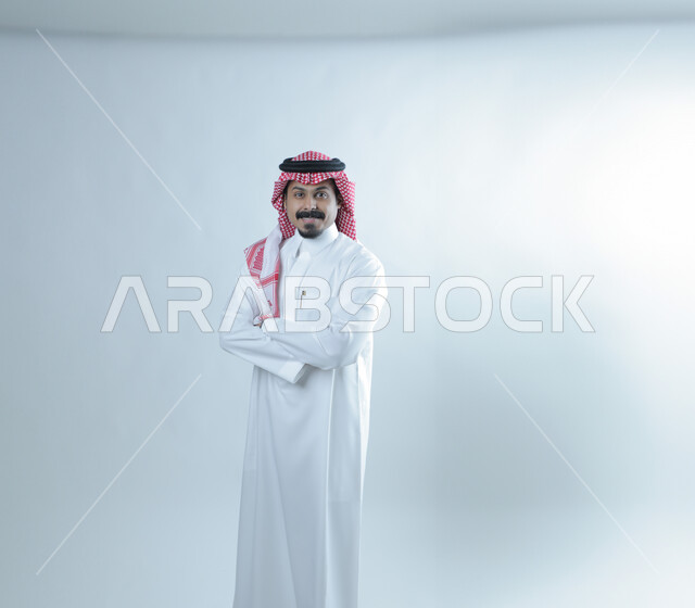 The concept of elegance and masculinity, expressions of cheerfulness and pleasure, looking at the camera with gestures of self-confidence, interest in external appearance, portrait of a Saudi Arabian Gulf man wearing traditional thobe and shemagh standing straight with crossed hands, white background