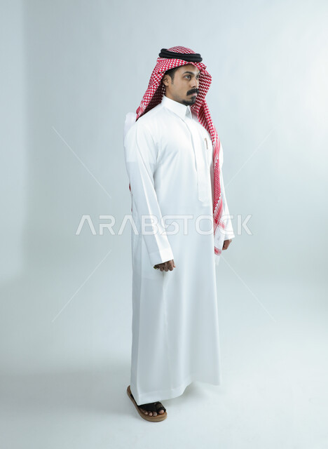 Caring about one's appearance, the concept of elegance and masculinity, expressions of cheerfulness and pleasure, looking at something with gestures of self-confidence, portrait of a Saudi Arabian Gulf man wearing a traditional thobe and shemagh standing straight, full body length, white background