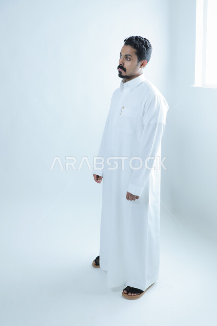 النظر إلى شيء ما بايماءات الثقة بالنفس، الإهتمام بالمظهر الخارجي، مفهوم الاناقة والرجولة بورتريه من الجانب لرجل عربي خليجي سعودي يرتدي الثوب التقليدي يقف باستقامة، الطول الكامل للجسم، خلفية بيضاء