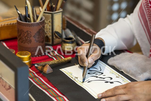فن الخط العربي، احتراف الكتابة بخط الثلث، صورة مقربة ليد رجل خطاط عربي خليجي سعودي يمسك بيده الريشة الخاصة ويكتب على لوحة أمامه، تعليم مهارات الرسم بالخطوط بشكل انسيابي، التمسك بالثقافة العربية الأصيلة في السعودية، نقش الكلمات وكتابتها بشكل فني هندسي مميز